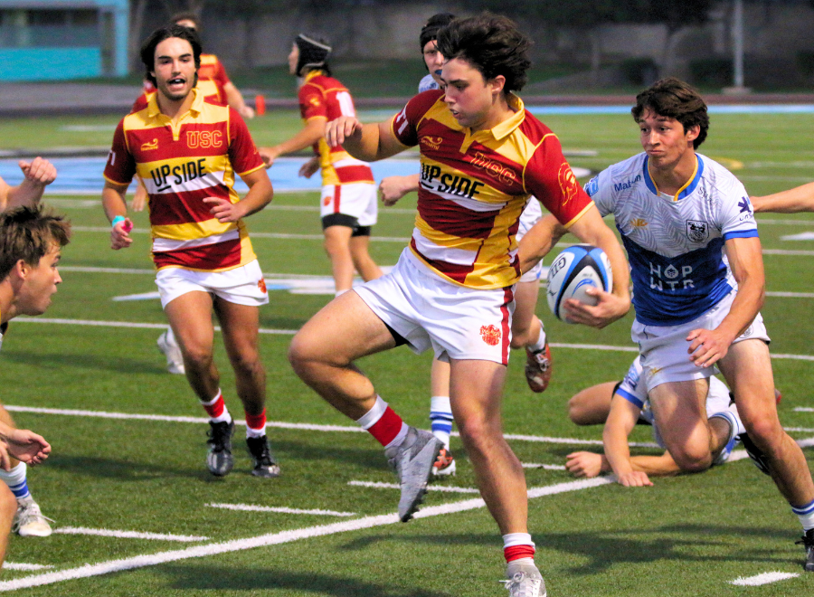 Rugby player sprinting across the field while clutching the ball, closely pursued by a defender attempting to tackle from behind.